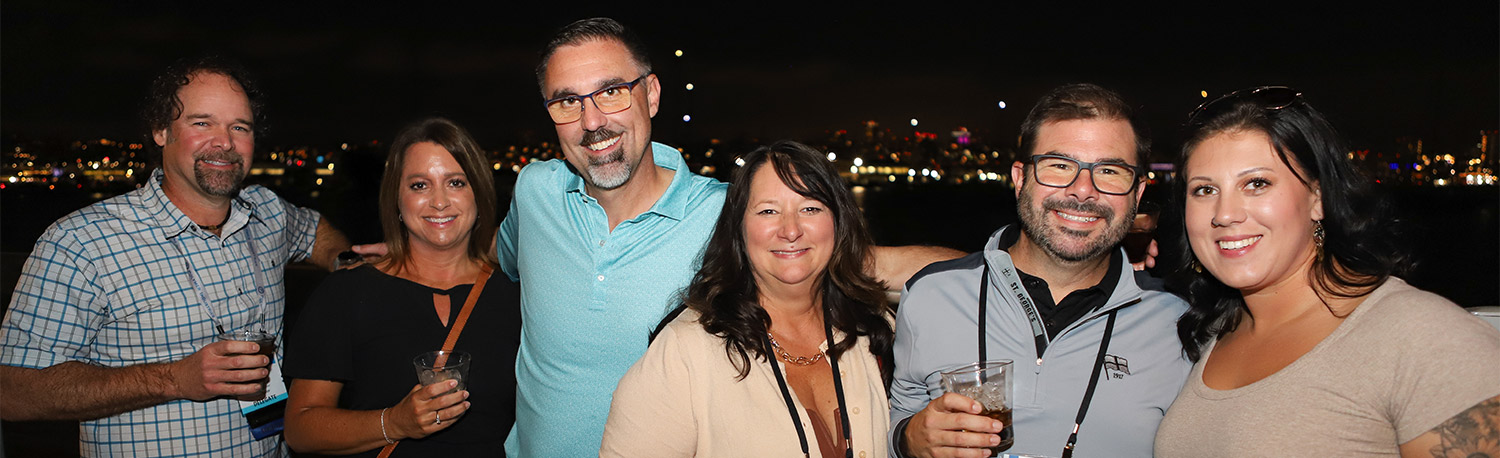 six attendees of the 67th IAPD Annual Convention closing party take a group photo on a balcony at Coasterra, lights from downtown San Diego shine along the horizon in the background