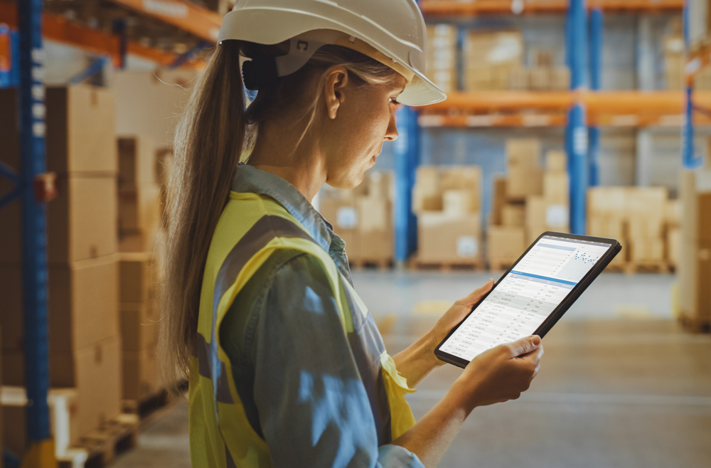 woman wearing a hardhat and neon yellow safety vest holding a tablet in a warehouse with boxes