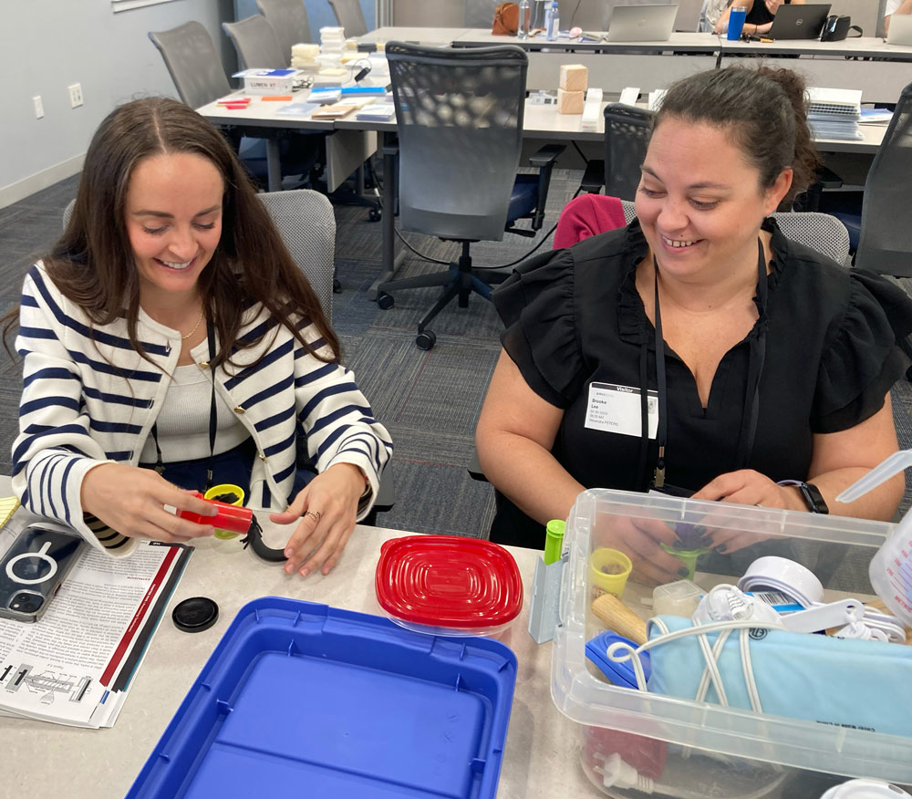 Two women seated at a table, engaged in conversation, with a box of plastic materials placed between them