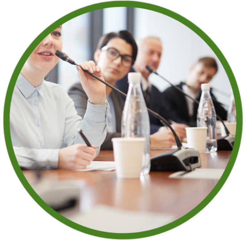 A cropped image shows four people sitting at a long wooden table, each with a microphone in front of them, suggesting they are on a panel or speaking at a conference.