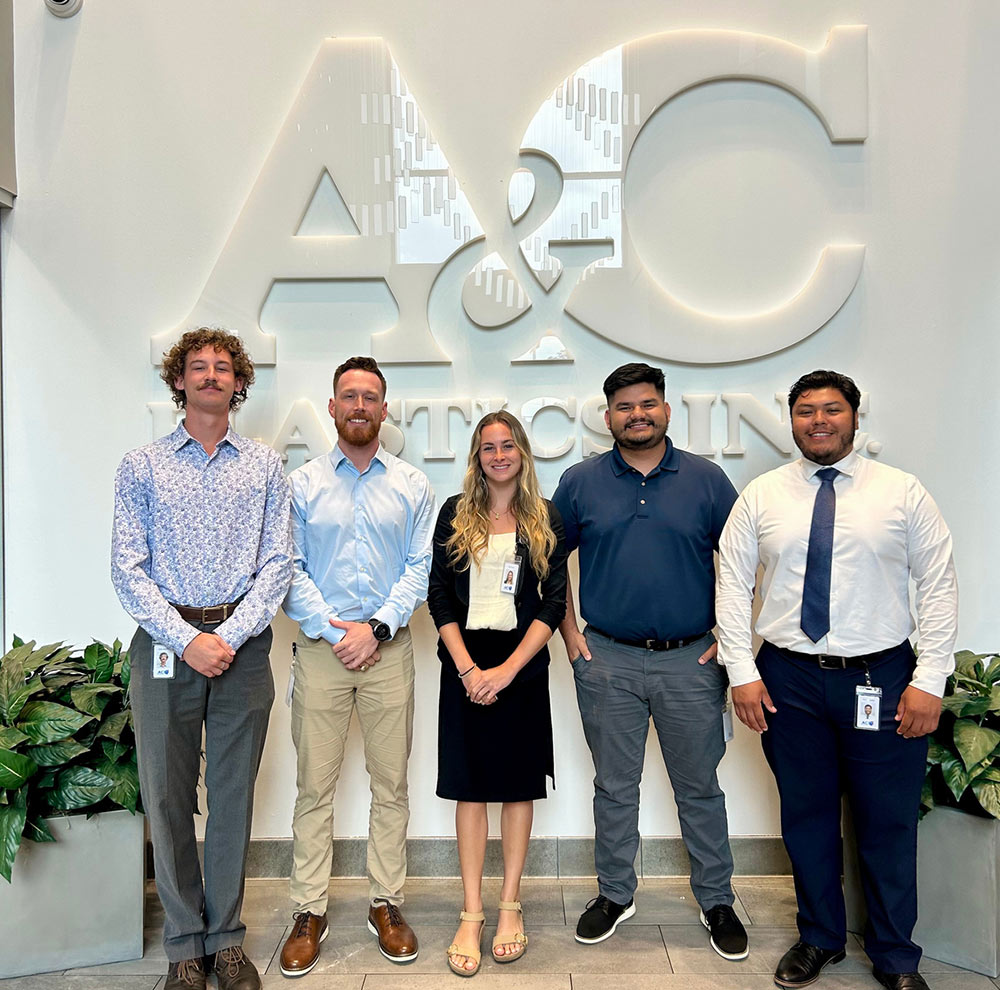 five A&C Plastics summer interns, four men and a woman take an indoor group photo in while standing in front of A&C Plastics large wall signage