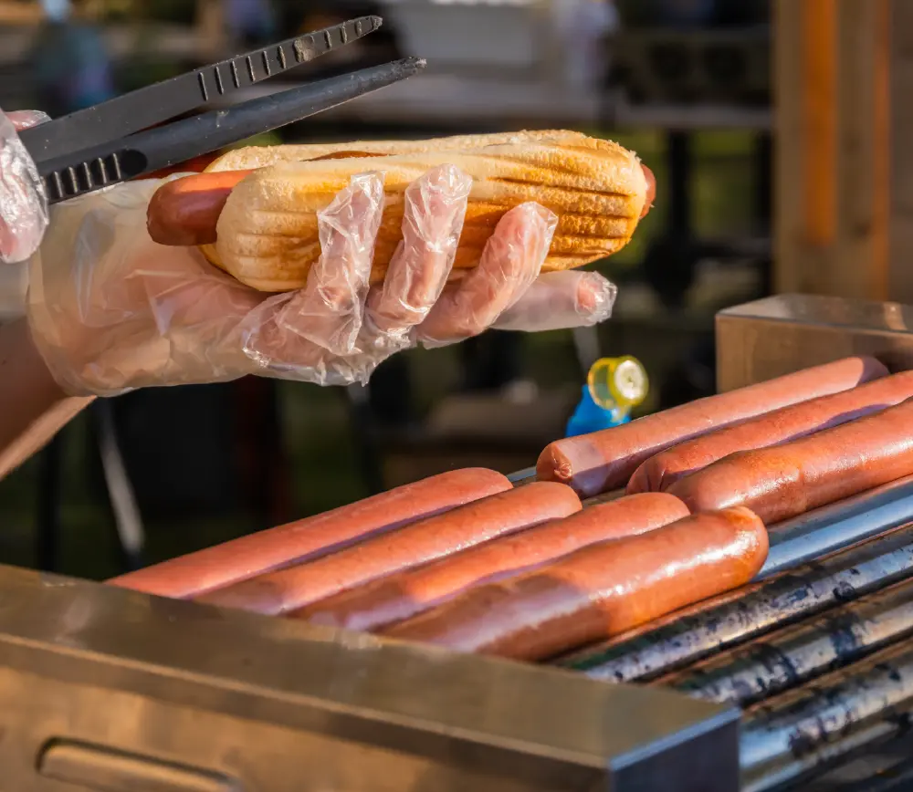  A pair of hands in clear plastic gloves holding a hot dog in a bun, with several other hot dogs on a grill in the foreground.