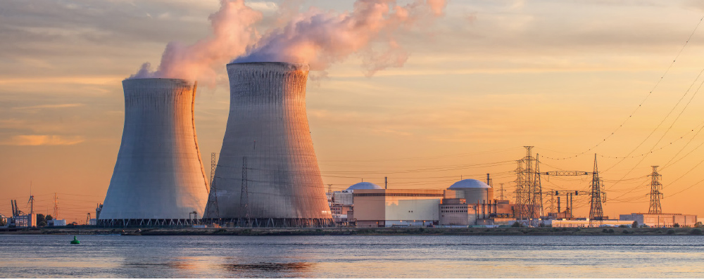The image shows a large industrial complex, likely a nuclear power plant, dominated by two massive cooling towers releasing plumes of steam against an orange and yellow sunset sky.