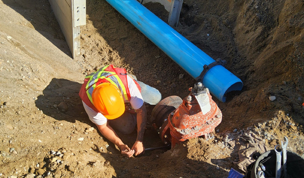 A construction worker in an orange hard hat and reflective safety vest is kneeling in a trench, working on a reddish-brown pipe fitting.
