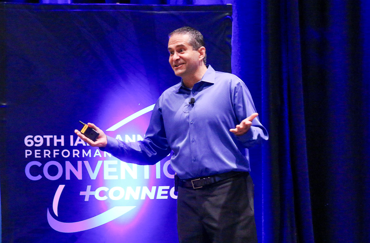 A speaker in a blue shirt gestures with open hands while presenting on stage. He holds a remote control in front of a banner for the 69th IAPD Annual Performance Convention + Connect against a dark blue curtain.