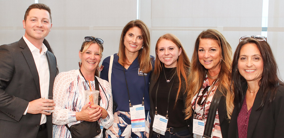 Six smiling attendees pose together in a hallway at the IAPD convention. They wear business casual attire and name badges, with one holding an iced coffee and another holding a beverage glass.