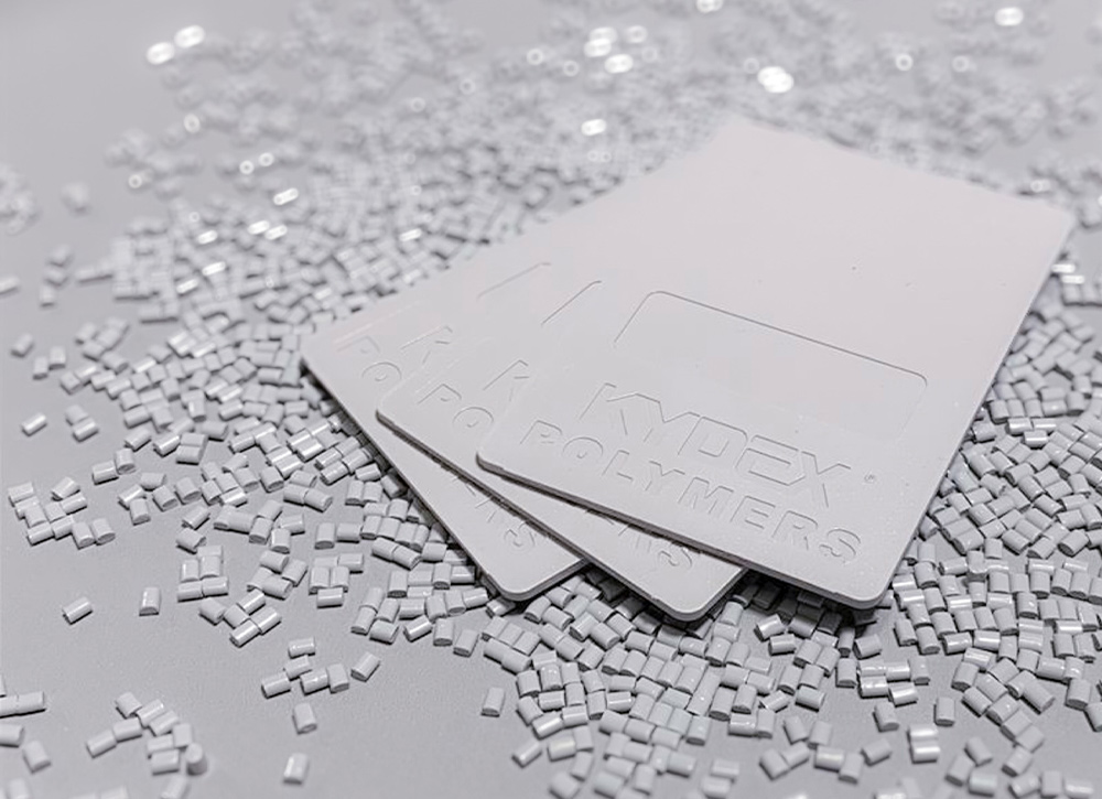 Three light grey KYDEX thermoplastic sheets with embossed logos rest atop a bed of small, matching injection molding resin pellets on a flat surface.