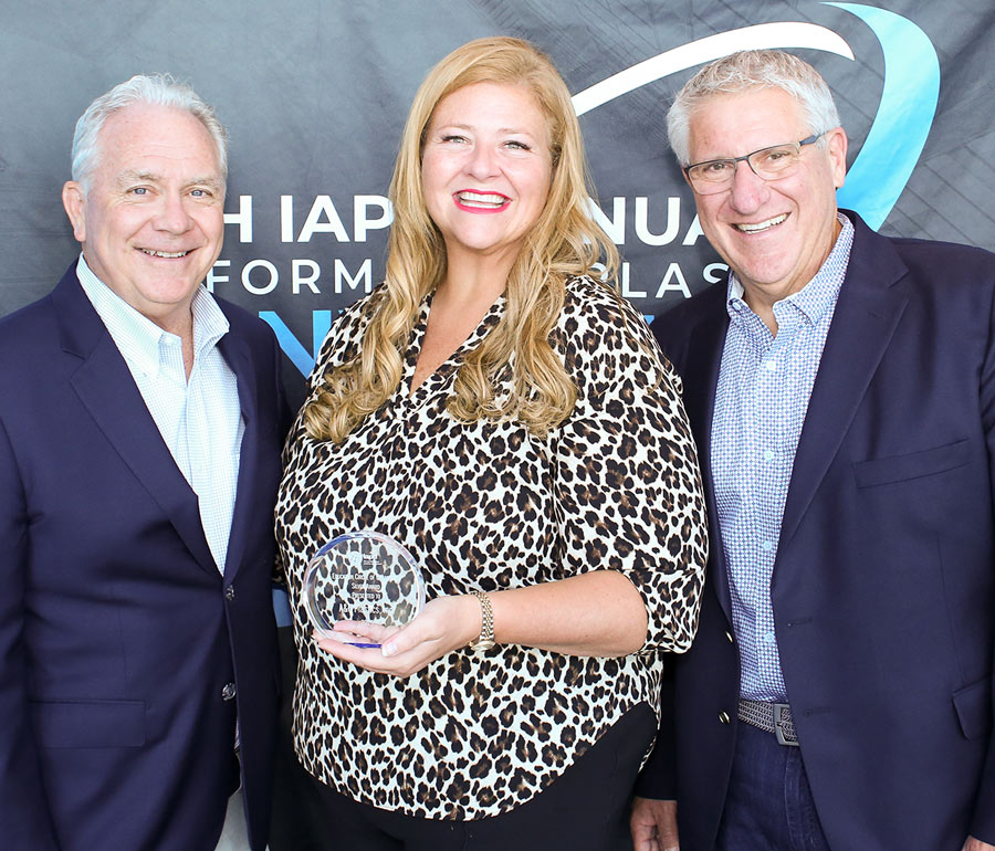 Katie Clapp of A&C Plastics, Inc. smiles between two presenters in front of a convention banner. She wears a leopard-print top and holds a clear, circular Silver Education Circle of Champions award plaque.