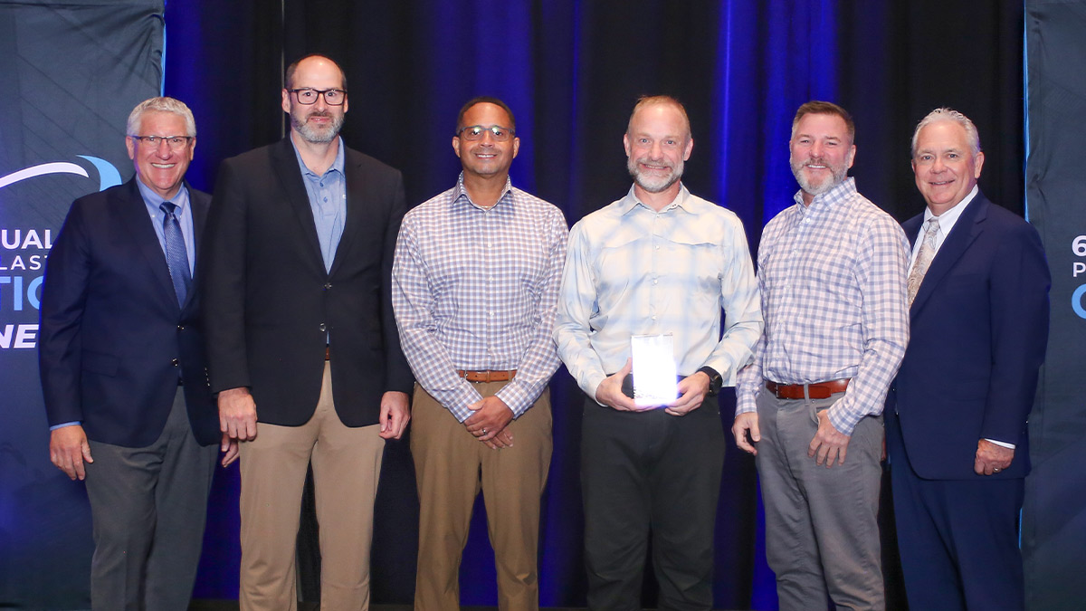 Six men stand in a row against a blue draped background. Four Ensinger representatives pose between two presenters in suits, with one recipient holding a clear Environmental Excellence award.