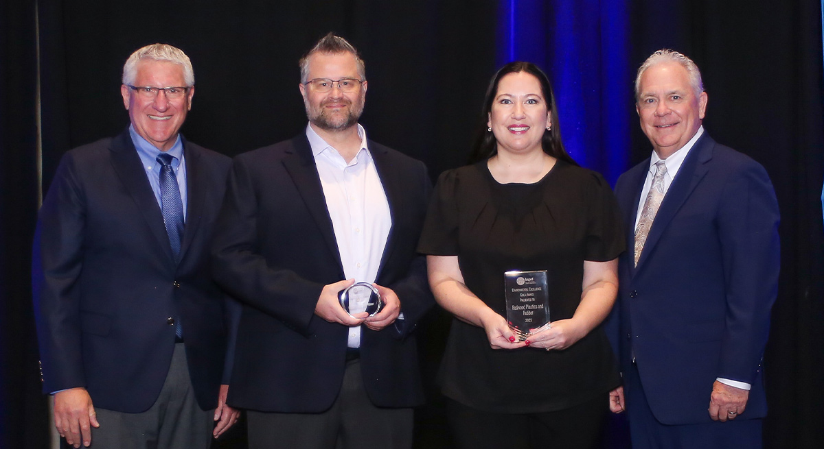 Two Redwood Plastics and Rubber representatives pose between two presenters against a blue backdrop. A man holds a circular award while a woman holds a rectangular Gold Level Environmental Excellence plaque.