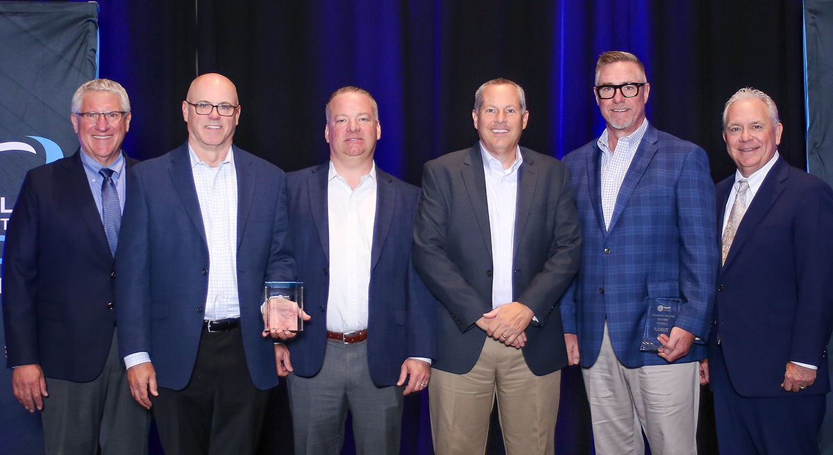 Four Plaskolite representatives stand between two presenters against a blue backdrop. Two men hold clear Silver Level Environmental Excellence award plaques.