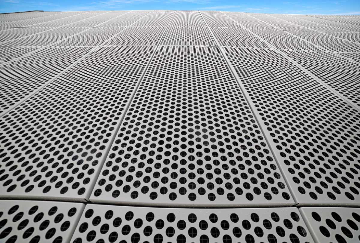 A low-angle perspective of a massive exterior building facade made of light gray perforated metal panels with a repeating circular hole pattern against a clear blue sky.