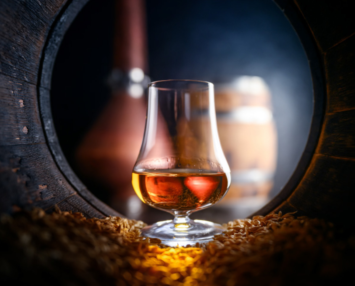 A glass of amber liquid stands in the center of a circular wooden barrel opening, resting on a bed of golden grain with a distillery pot still in the background.