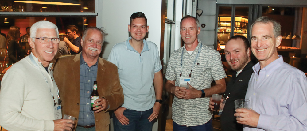 Six men socialise with drinks during a nighttime reception at the performance plastics convention, posing for a group photo in a well-lit indoor lounge area.