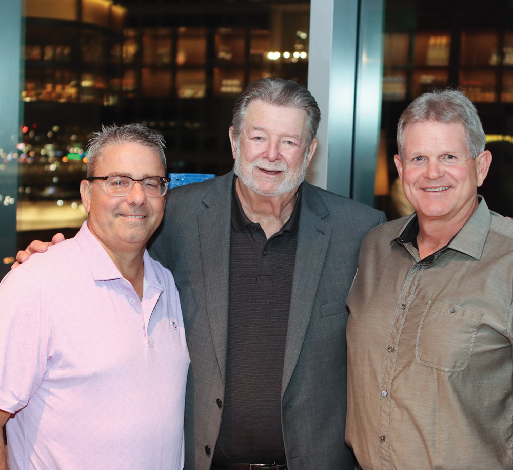 Three men stand close together for a friendly portrait at a nighttime event, featuring views of city buildings through the windows behind them.