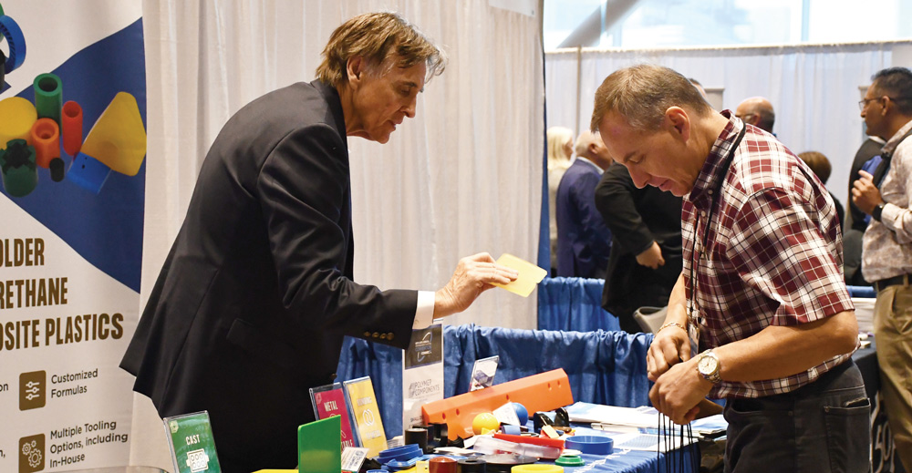 A man in a suit displays a yellow plastic sample to an attendee at a trade show booth featuring various colorful polymer components and molded urethane products.