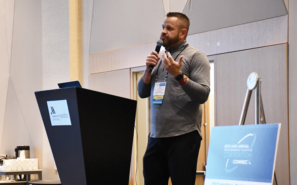 A speaker in a grey pullover gestures while holding a microphone.