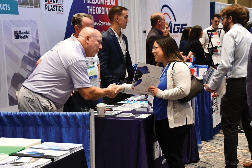 An exhibitor shakes hands with a woman holding a brochure at a professional trade show booth. Several attendees converse in the background of the convention hall.