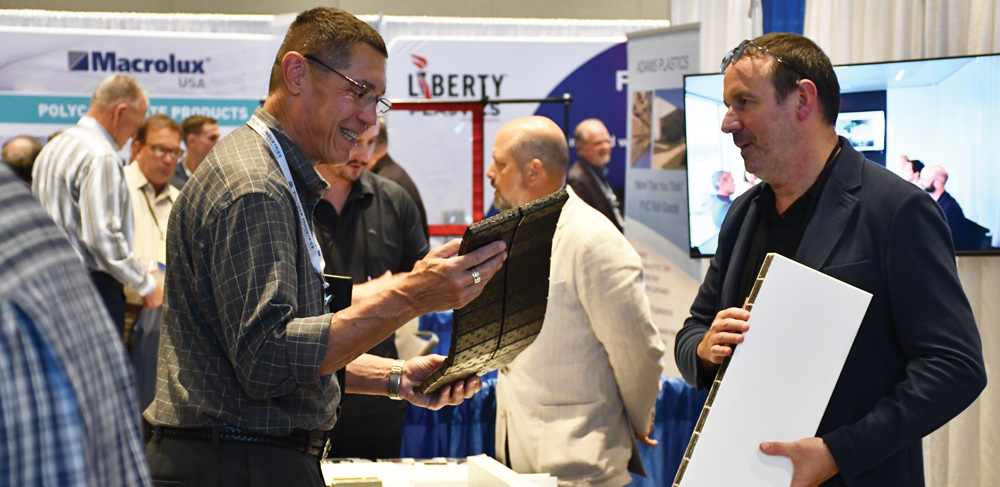 Two men at a trade show examine different industrial plastic samples, including a textured black flexible sheet and a thick white rectangular board.