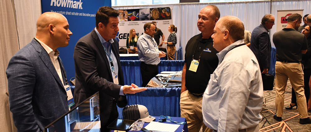 A group of professionals in business attire engage in a discussion at a Rowmark brand booth during a plastics industry trade show and convention.