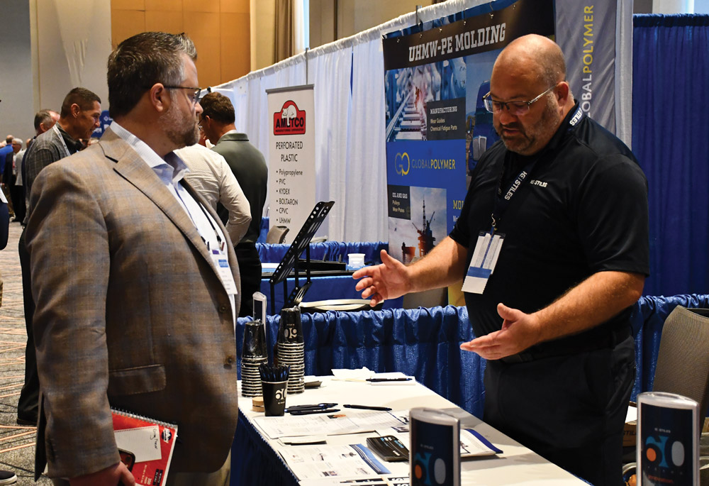 An exhibitor at a booth gestures while talking to a man in a blazer at a professional industry trade show.