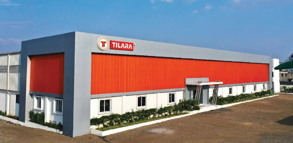 Exterior view of a large, modern industrial building with a grey and vibrant orange facade. The Tilara logo is prominently displayed on the upper left corner of the grey section above a row of windows and a small entrance canopy.