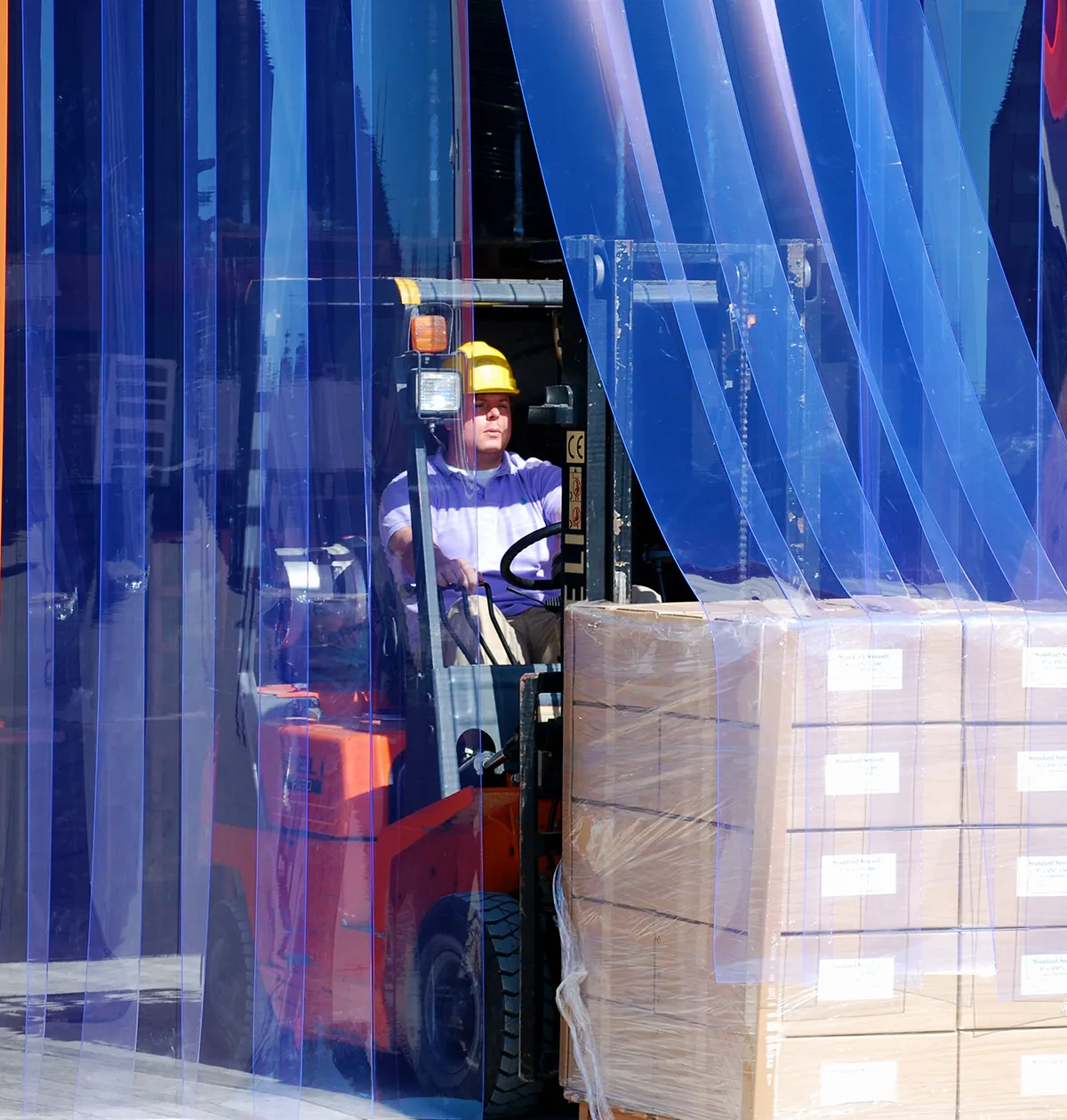 A warehouse worker in a yellow hard hat operating an orange forklift, driving through a protective blue PVC strip curtain while carrying a pallet of stacked cardboard boxes