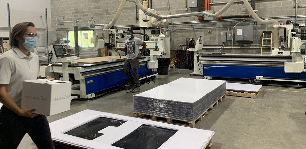 A busy plastic fabrication facility featuring workers in face masks operating large CNC routing machines to process white plastic sheets and manage finished product inventory