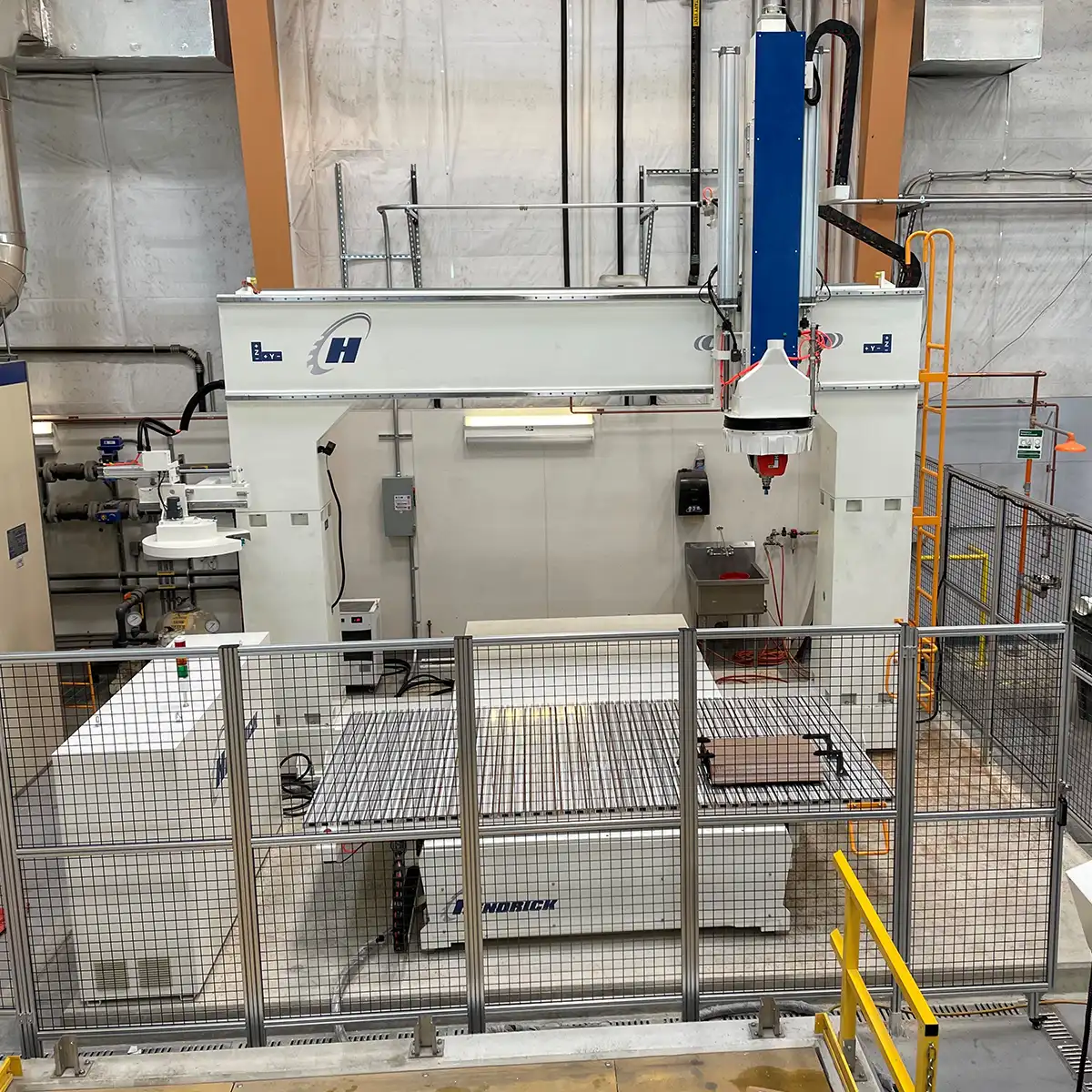 A large, industrial Hendrick CNC router with a blue and white gantry, enclosed in a safety cage within a manufacturing facility for high-precision plastic and wood fabrication