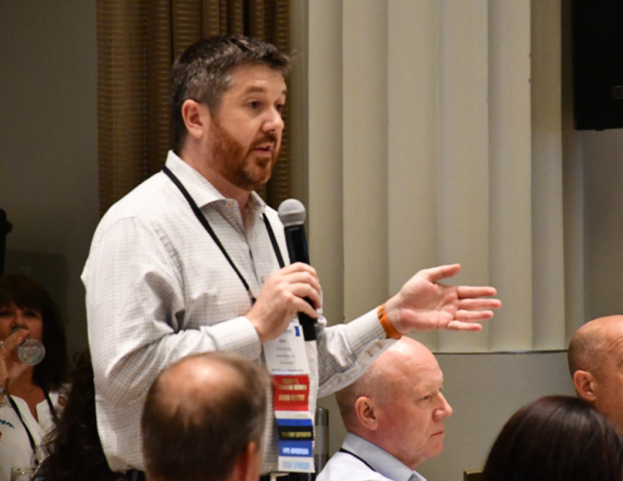 a man holding a microphone and wearing a lanyard displaying information addresses a seated audience in a forum like setting
