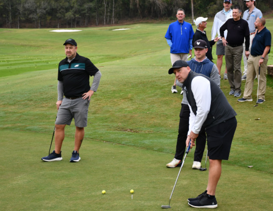 three men stand on a golf green as a small group of others mingle near by