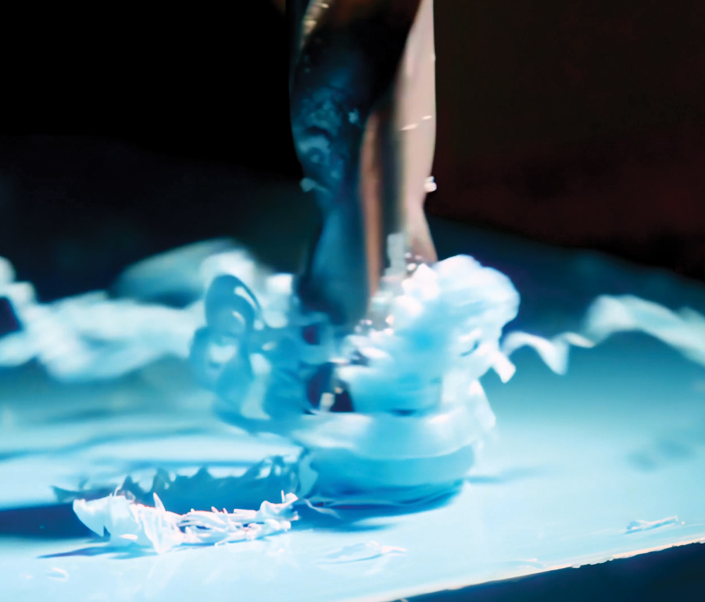 Extreme close-up of a drill bit penetrating a blue composite material, showing the rapid generation of fine, light-colored shavings and debris at the point of contact.