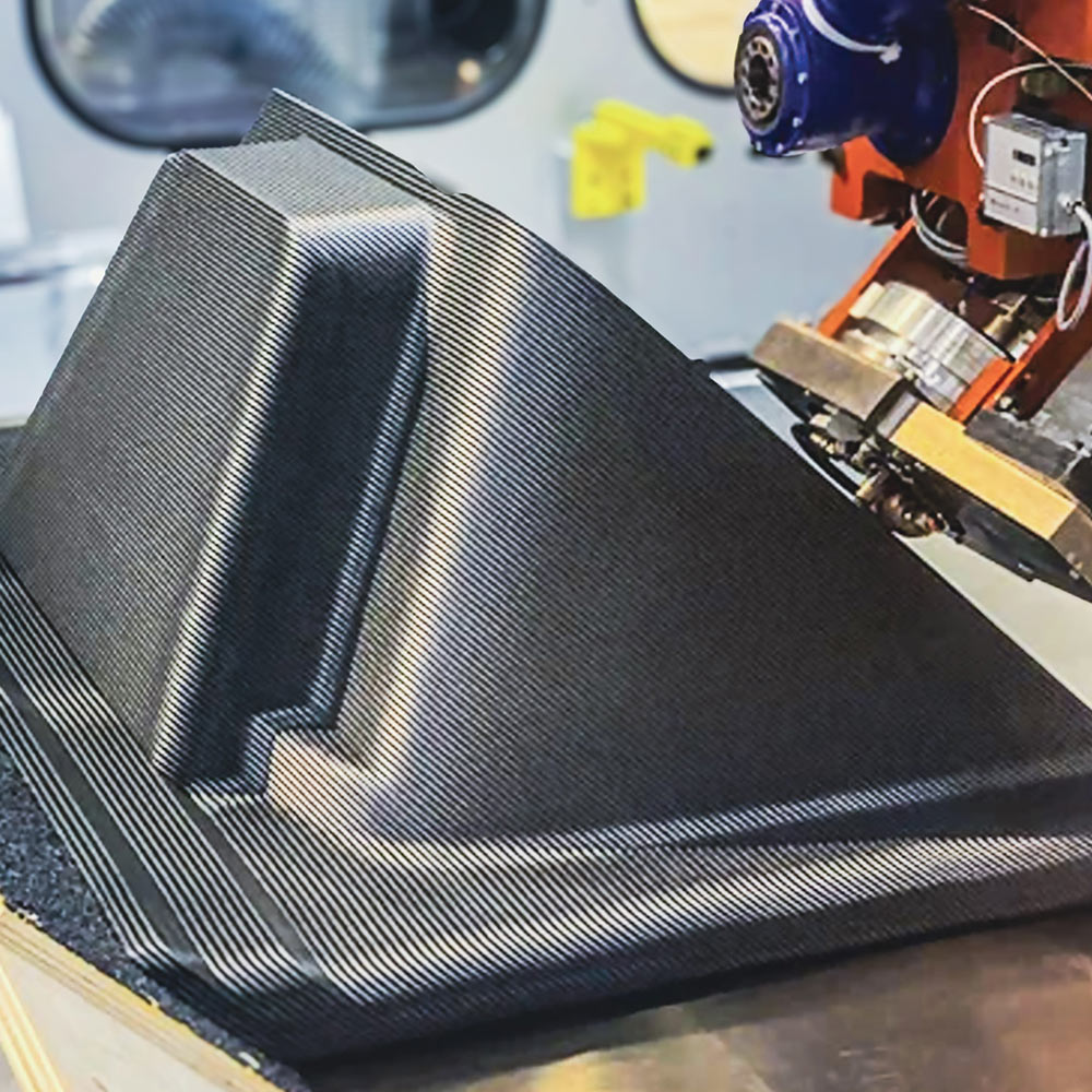 Close-up of a large-scale 3D printer extruding layers of carbon fiber reinforced polymer to create a complex, angled industrial part with a distinct ribbed surface texture.