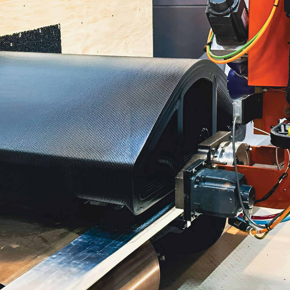 A robotic additive manufacturing head deposits thick, precise layers of black composite material onto a curved, hollow cylindrical structure in a controlled factory setting.