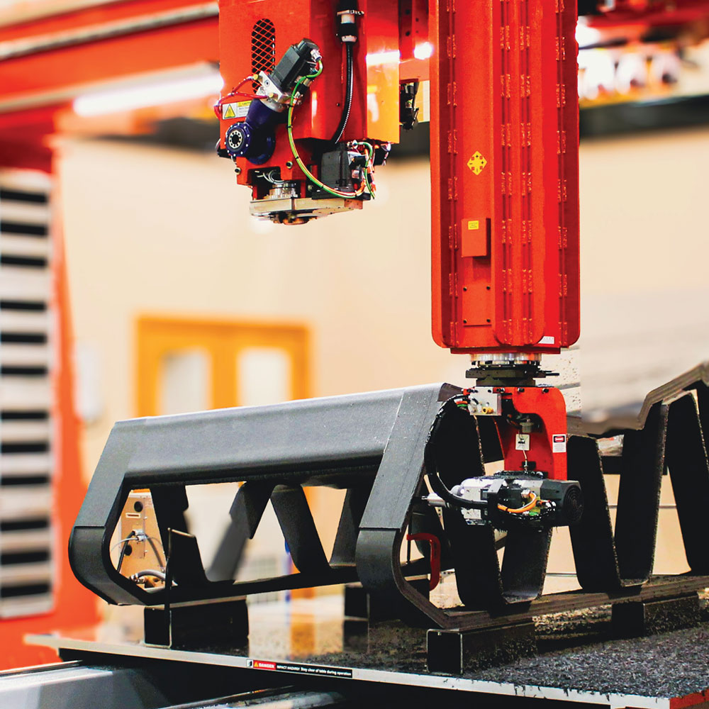 A large red gantry-style industrial robot uses an additive manufacturing nozzle to print a massive, structural black composite component on a flat production bed.