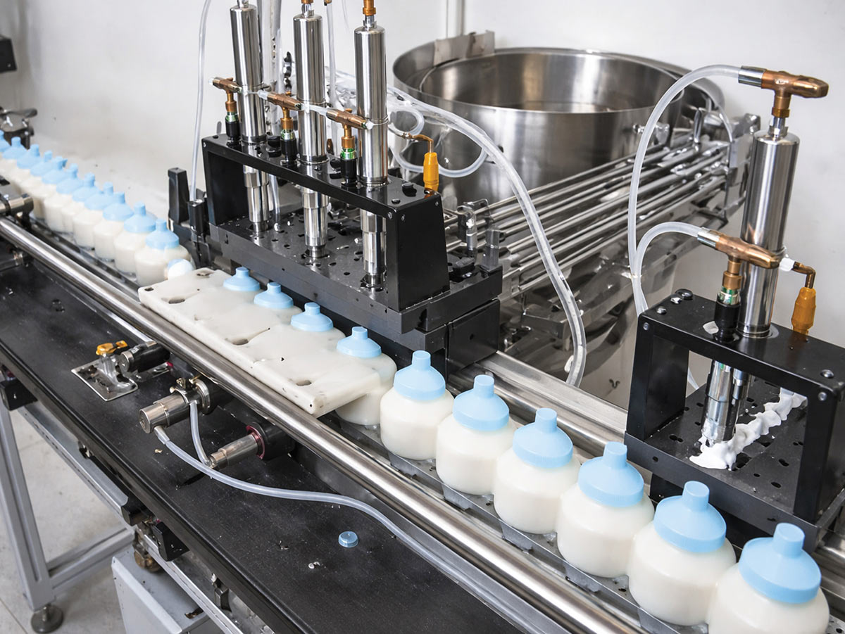 Automated production line filling small white bottles with light blue caps using stainless steel nozzles and clear tubing in a manufacturing facility