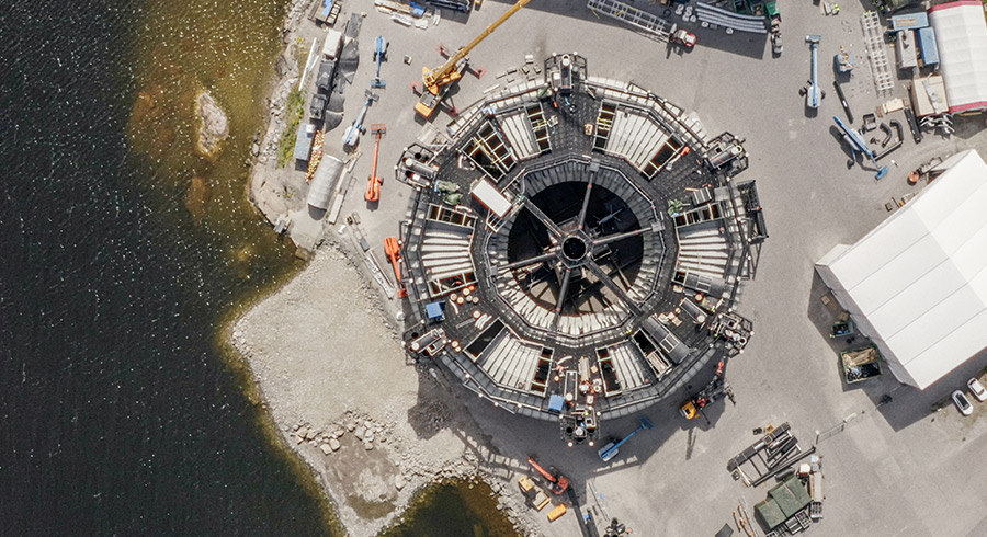 Aerial view of a large, octagonal thermoplastic structure, known as a Marine Donut, under construction in a shipyard near the water.