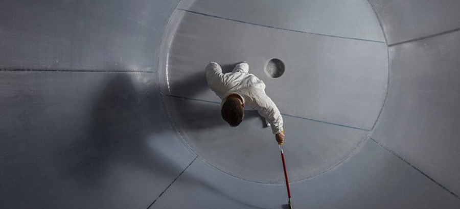 Overhead view of a person in a white cleanroom suit working on the interior surface of a large, circular grey storage tank liner.