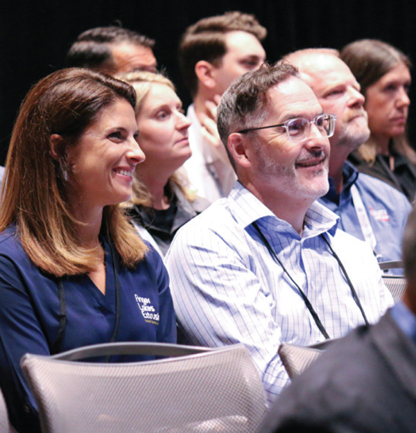 A man and woman smile while sitting in an audience at a professional event.