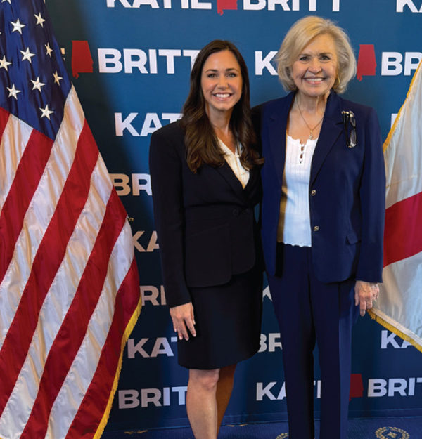 Two women stand together smiling in front of an American flag and a backdrop featuring the name Katie Britt.