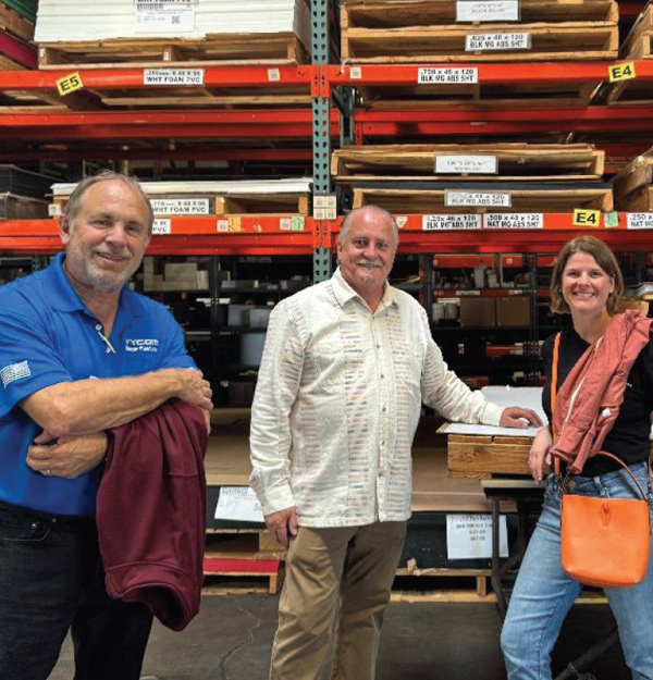 Three people stand together in a warehouse in front of high industrial shelving filled with stacked pallets of plastic sheet materials.