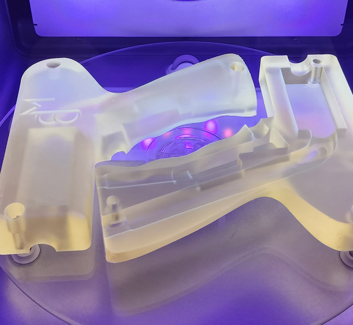 Two translucent white stereolithography (SLA) plastic components resting on a platform inside a curing chamber under purple light.