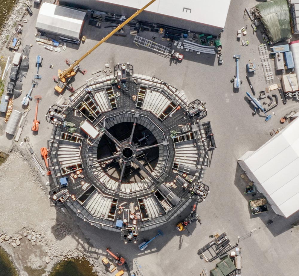 Aerial view of a large, octagonal thermoplastic structure under construction in an industrial yard, featuring a central support frame and surrounded by construction equipment and cranes.