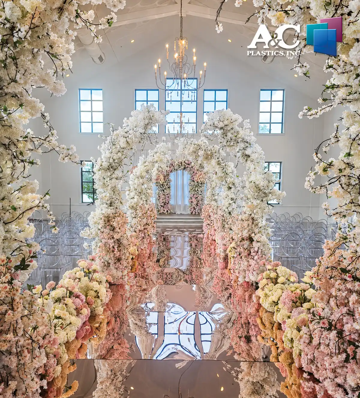 An elegant wedding venue featuring a highly reflective mirrored aisle beneath grand arches of white and pink flowers. Rows of clear acrylic chairs flank the aisle. The A&C Plastics, Inc. logo is in the top right corner.