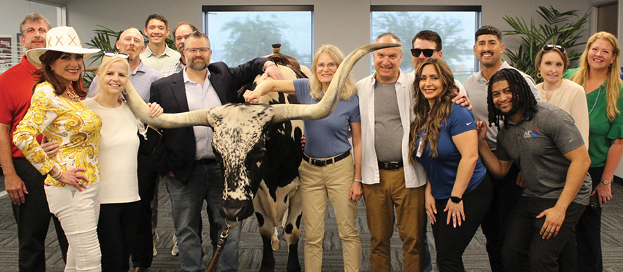 A group of attendees pose indoors around a black and white longhorn bull with wide horns at the A&C Plastics, Inc. headquarters.