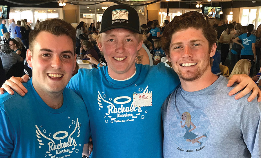 Three men pose shoulder-to-shoulder. Two wear bright blue T-shirts reading “Rachael's Warriors”, and one wears a light blue shirt with a Rachael Warrior Foundation graphic.