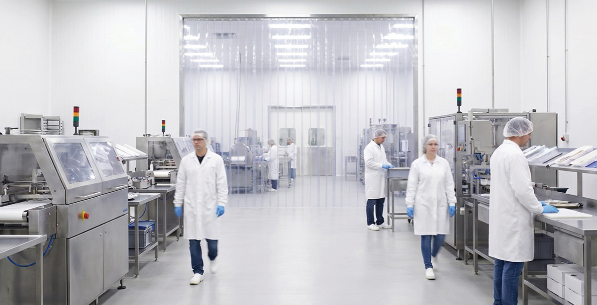 Workers in white lab coats and hairnets operating machinery in a clean manufacturing facility with a large clear plastic strip curtain doorway.