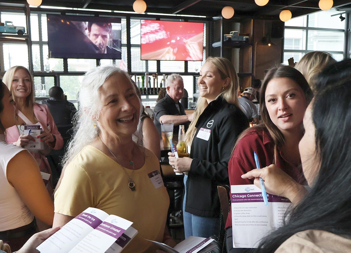 A group of women wearing name tags smiling and conversing while holding icebreaker activity sheets during a professional networking event.