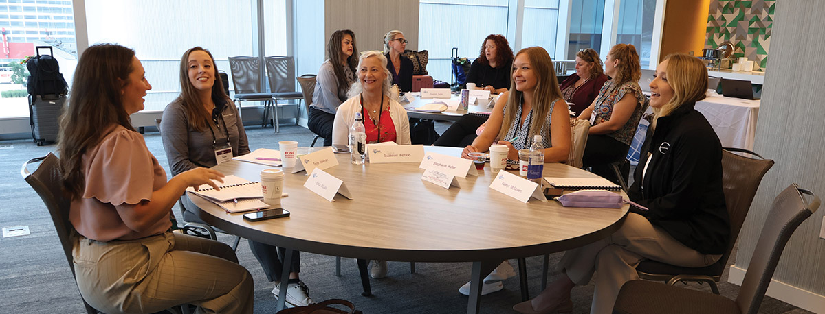 Women seated around a large round table with name placards and notebooks, participating in a group discussion during a conference session.