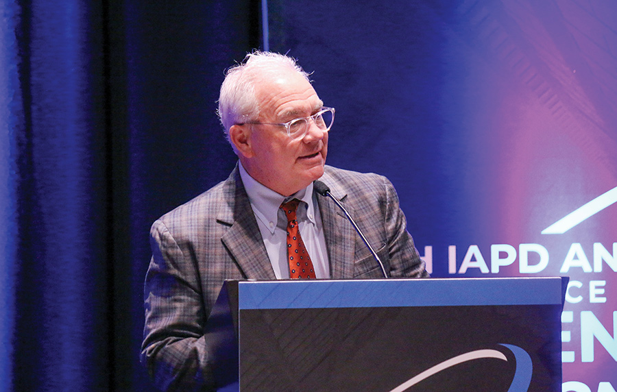 A man wearing glasses and a suit speaking into a microphone at a podium during an IAPD event.
